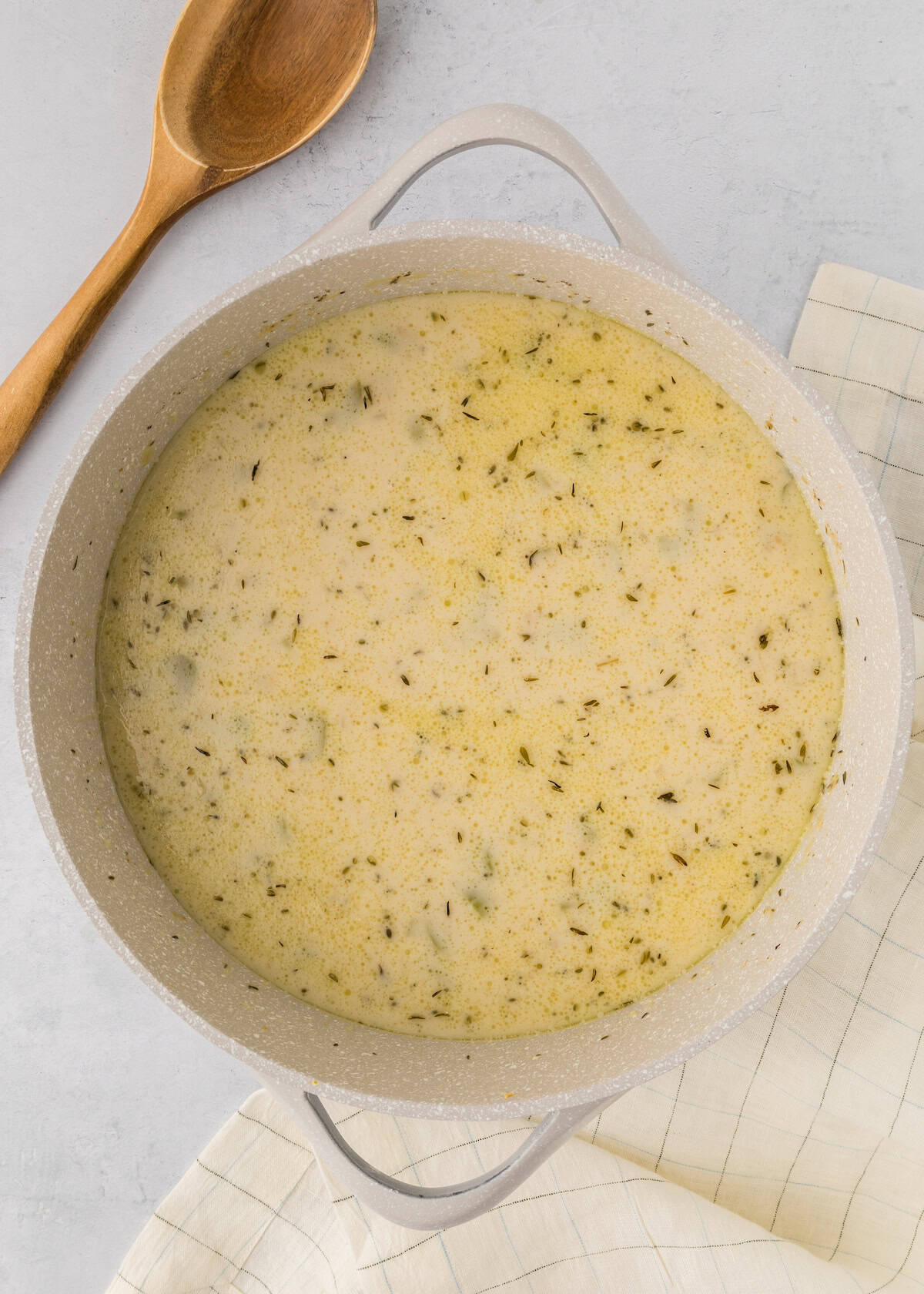 A pot of creamy soup with herbs next to a wooden spoon and a checkered cloth.