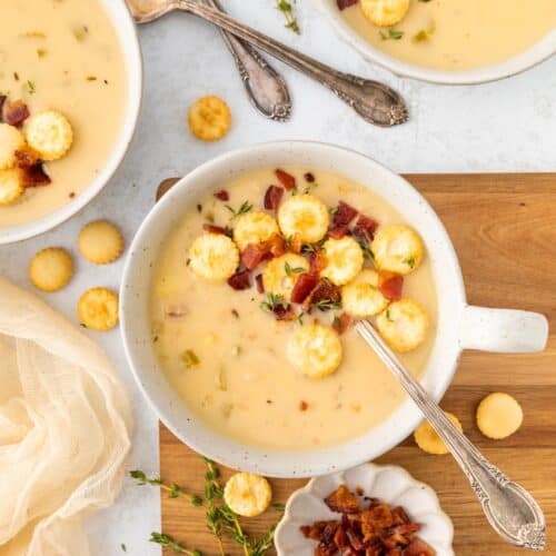 Creamy soup with crackers, bacon bits, and herbs in bowls, served with spoons on a wooden board.