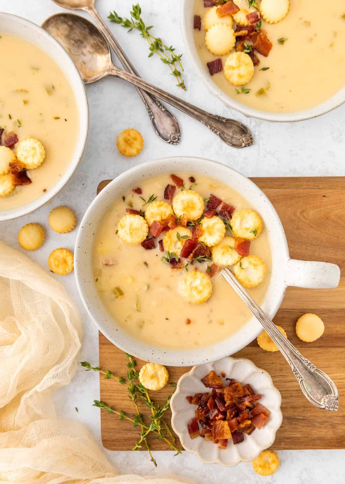Creamy soup with crackers, bacon bits, and herbs in bowls, served with spoons on a wooden board.