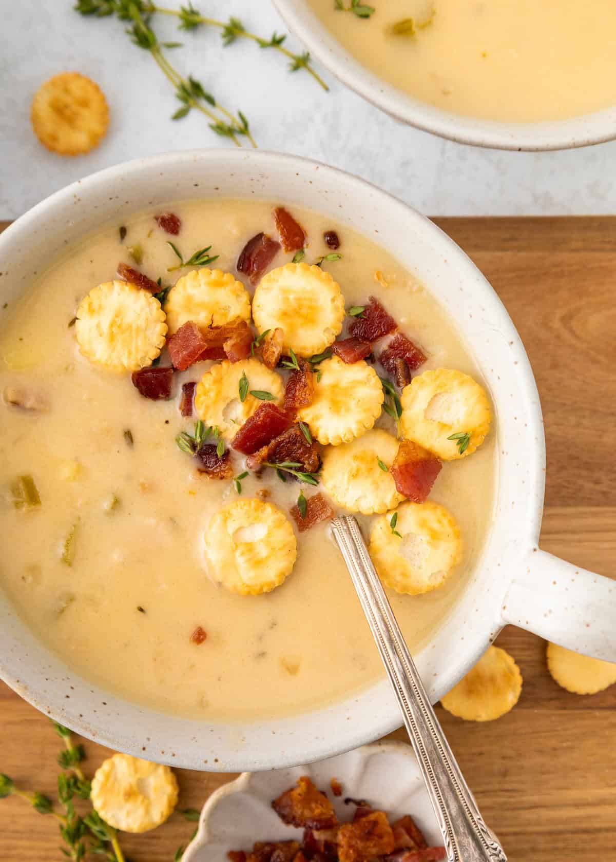 Creamy soup topped with oyster crackers, bacon, and herbs in a white bowl with a spoon.