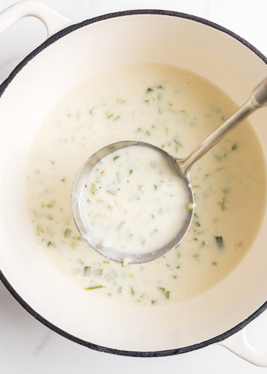 White soup with herbs in a cream-colored pot, being served with a metal ladle.