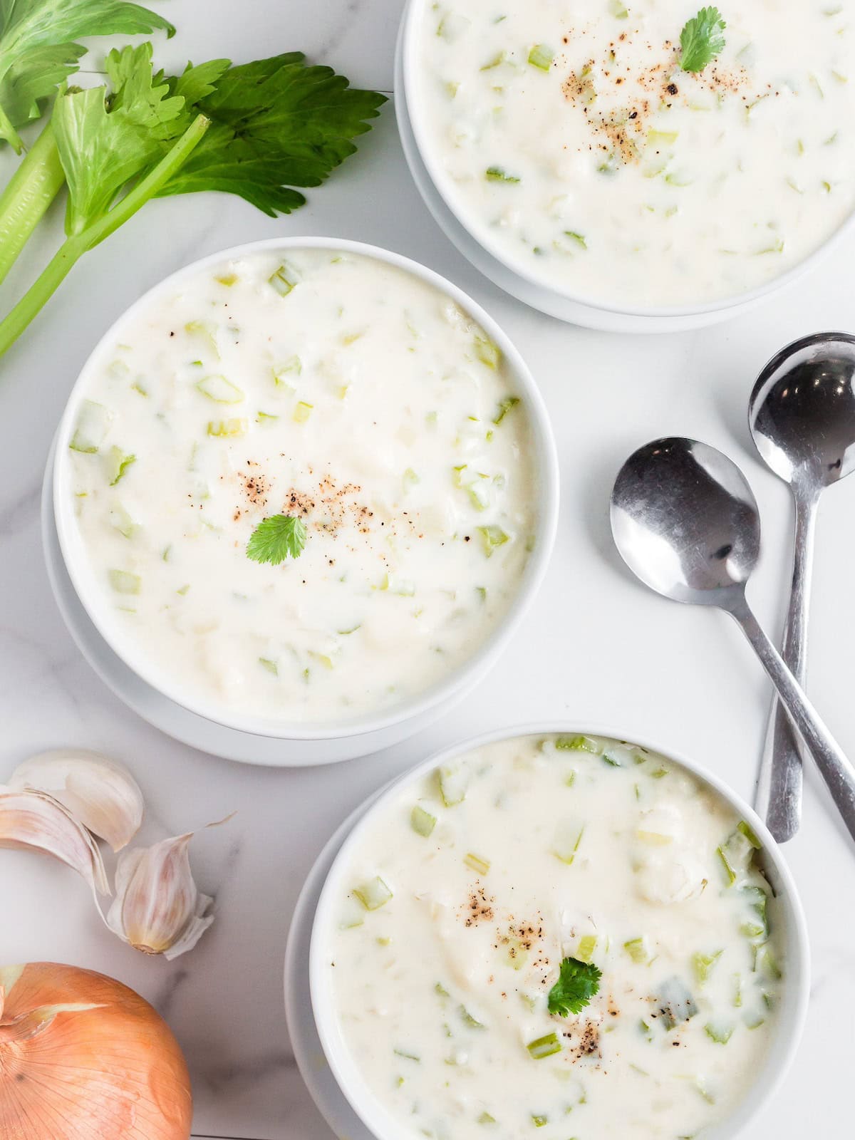 Three bowls of creamy soup with herbs, next to spoons, celery, onion, and garlic on a table.