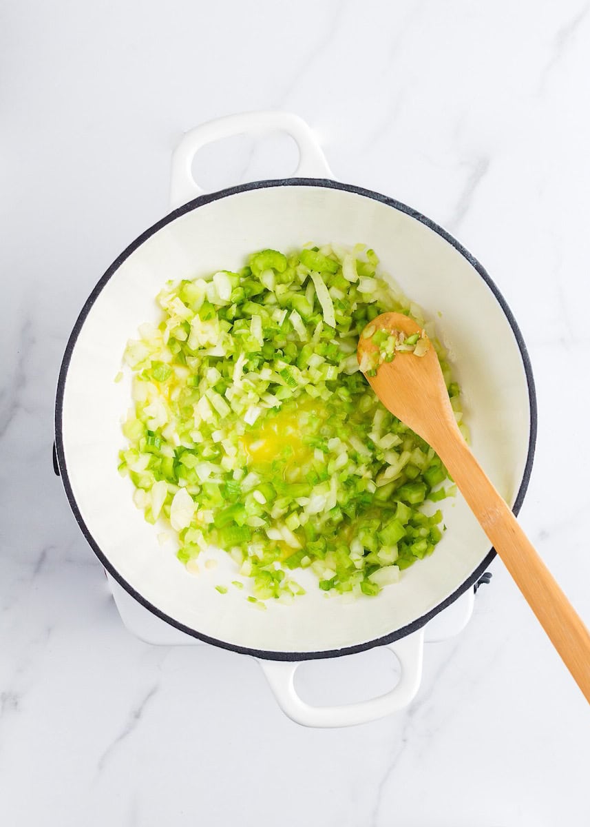 Chopped onions and celery being sautéed in a white pot with a wooden spoon.