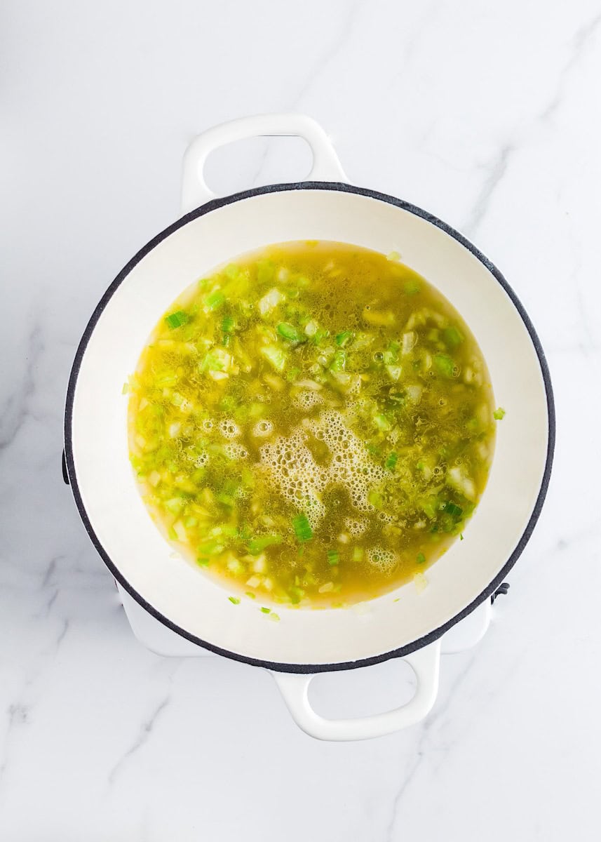 White pot with bubbling soup, containing chopped green vegetables and onions on a marble surface.