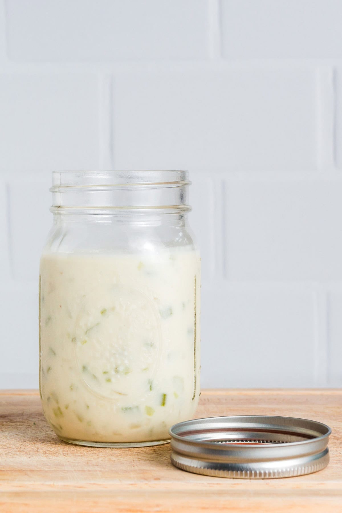 A glass jar of creamy dressing with herbs, sitting on a wooden surface beside its lid.