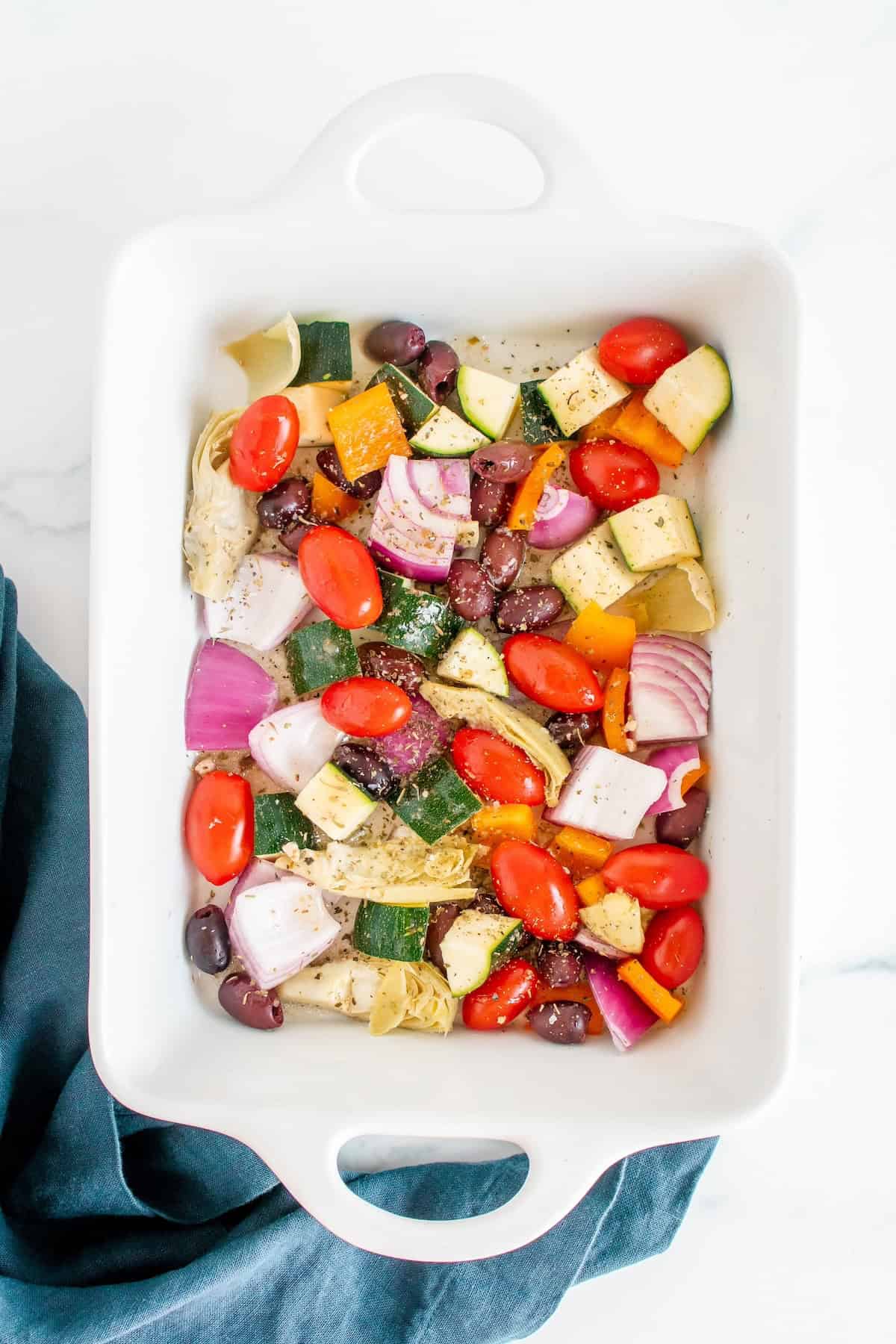 Chopped vegetables and olives in a white baking dish on a marble surface.