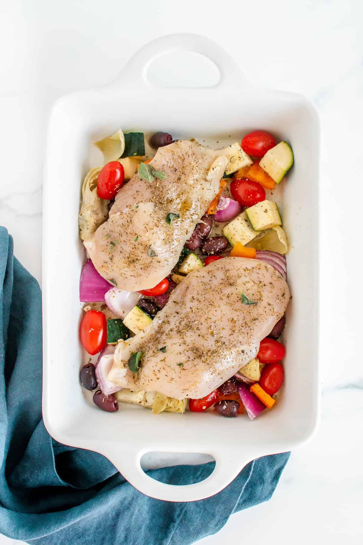 Raw chicken breasts on top of chopped vegetables in a white baking dish, ready to cook.