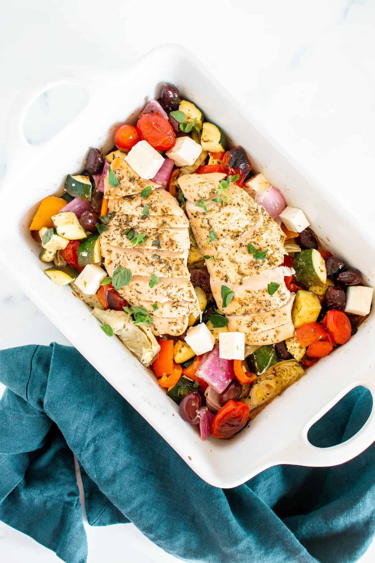 Baked chicken breasts with colorful vegetables, olives, and feta in a white casserole dish.
