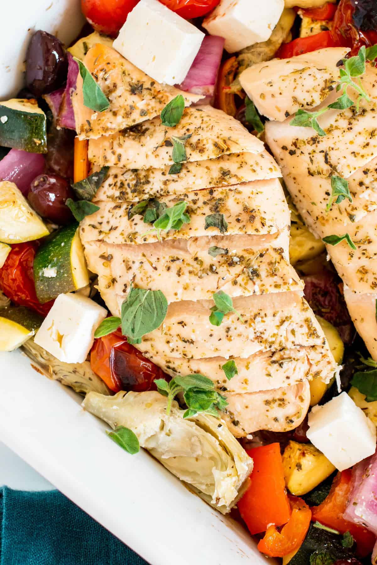 Sliced baked chicken breast with herbs, surrounded by colorful vegetables and feta cheese.