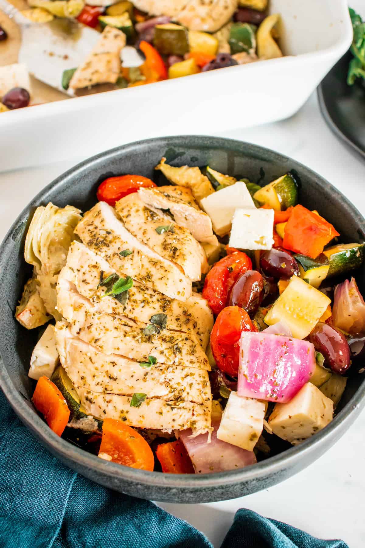 Sliced herb chicken with roasted vegetables and feta cheese in a black bowl.