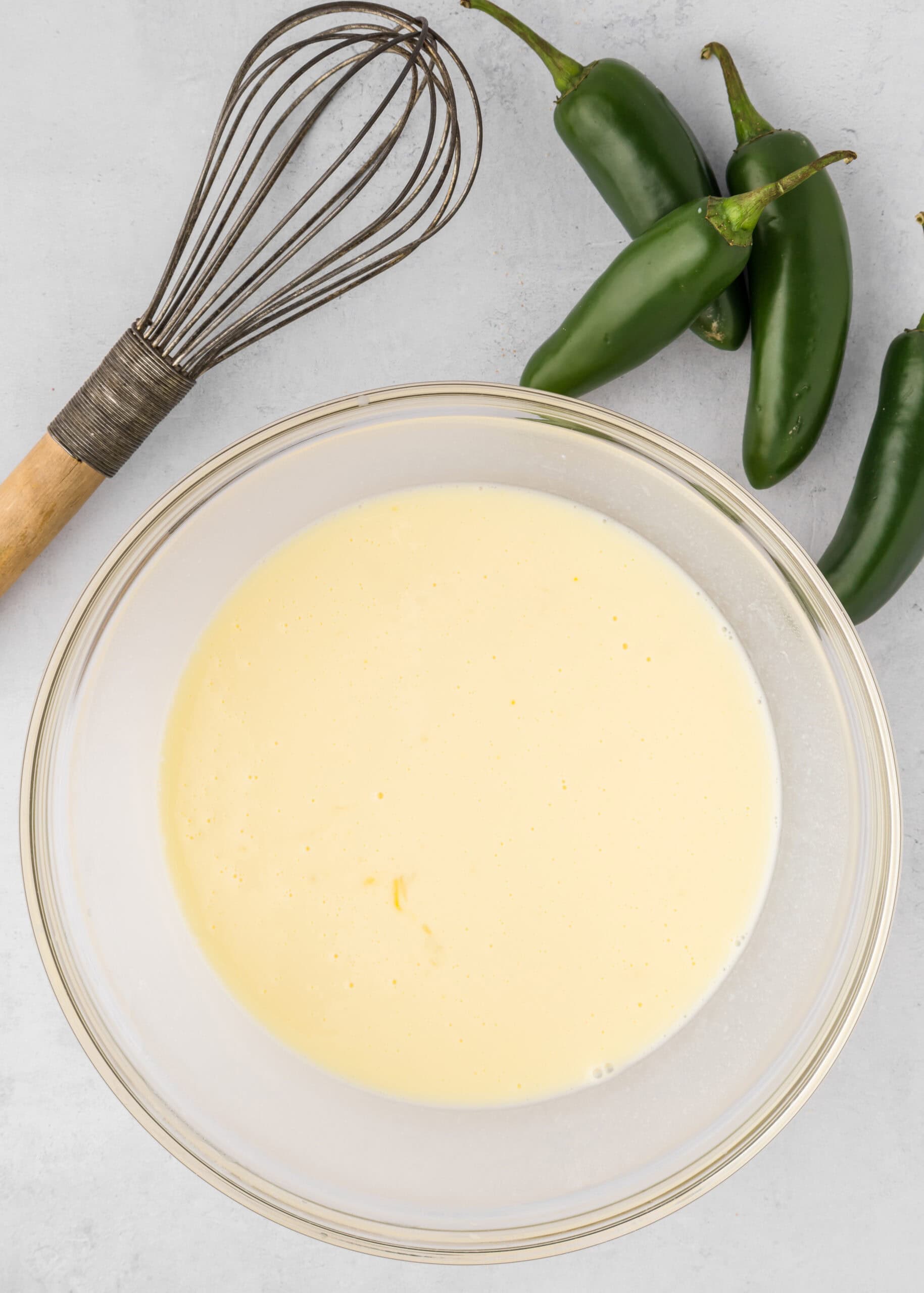 A bowl of creamy Jalapeño Cheddar Cornbread batter sits beside a whisk and fresh jalapeños.