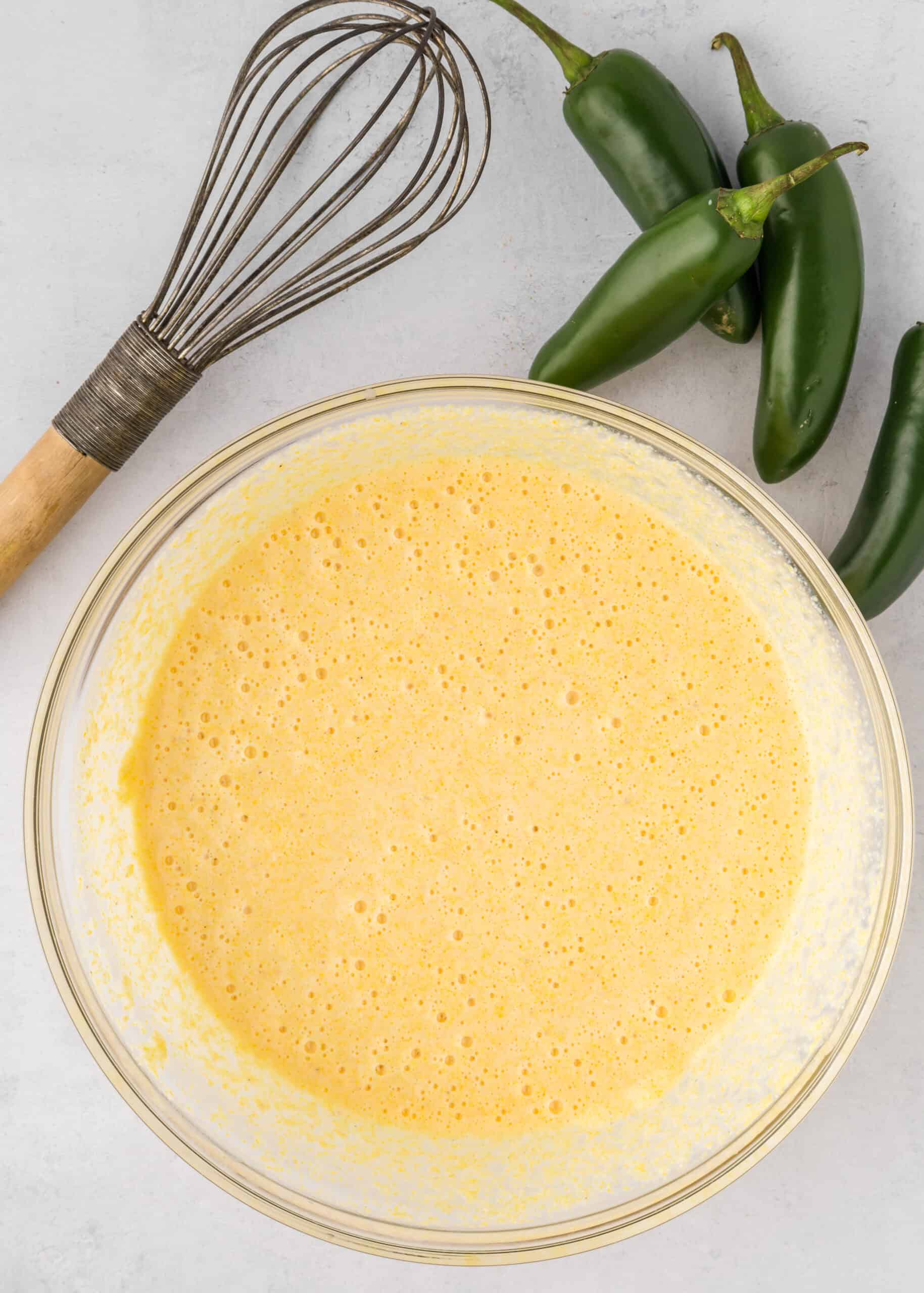 Bowl of Jalapeño Cheddar Cornbread batter with a whisk and fresh jalapeños on a light surface.