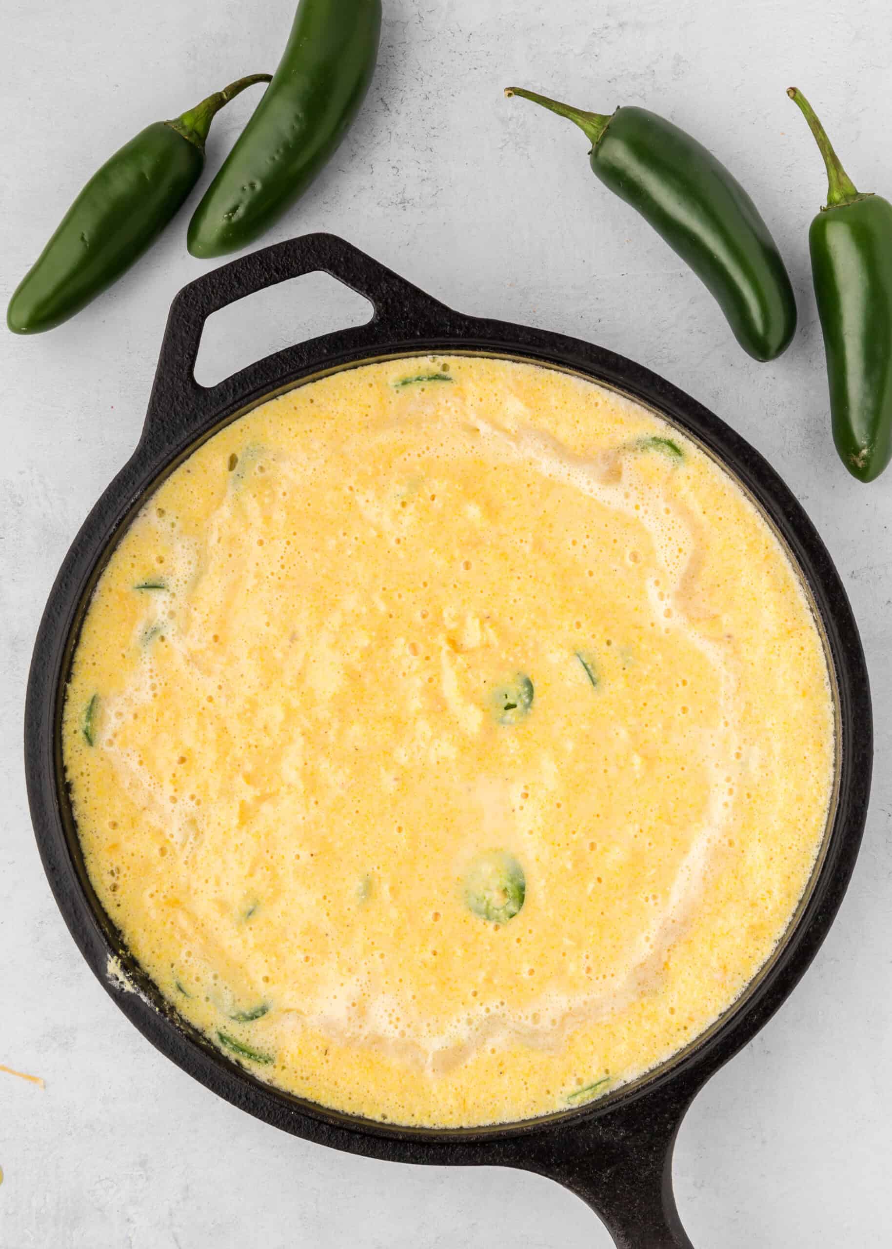 A cast iron skillet with Jalapeño Cheddar Cornbread batter, next to fresh jalapeños.