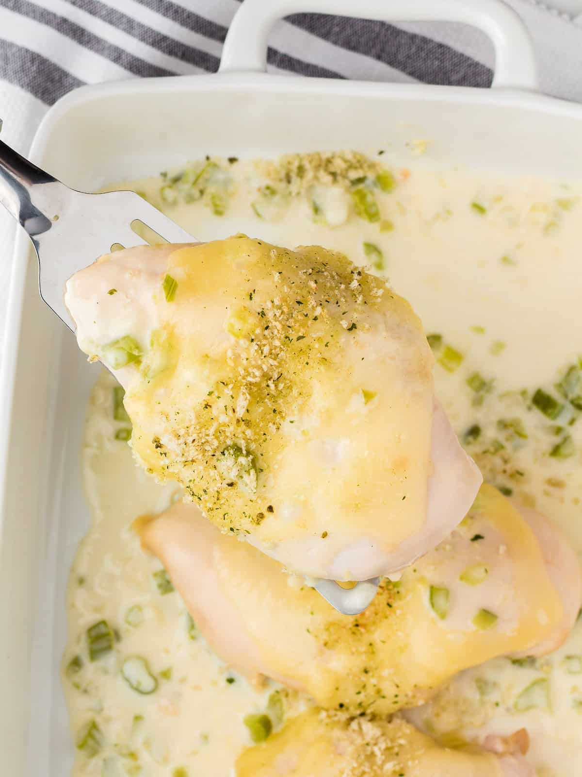 A spatula lifts cheesy baked chicken from a creamy casserole dish with green onions.