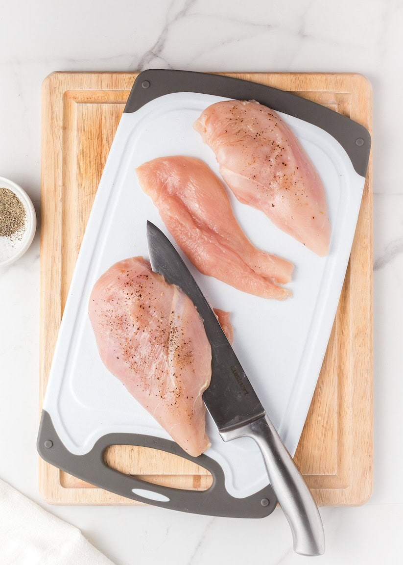 Raw chicken breasts with pepper on a cutting board, knife placed on top, next to pepper shaker.