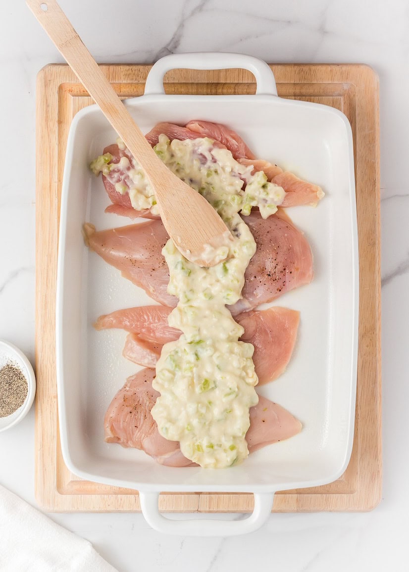 Raw chicken breasts in a baking dish topped with creamy mixture, being spread with a spatula.