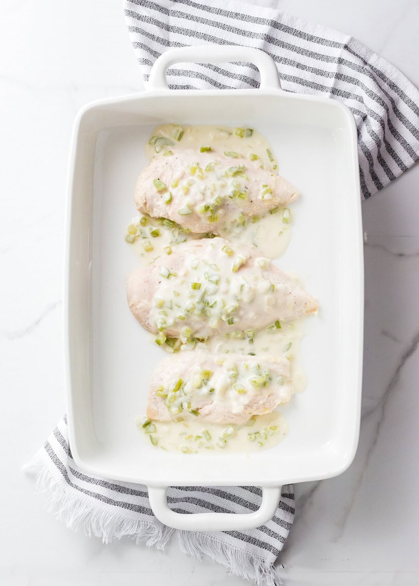 Three baked chicken breasts with creamy sauce in a white baking dish on a striped towel.