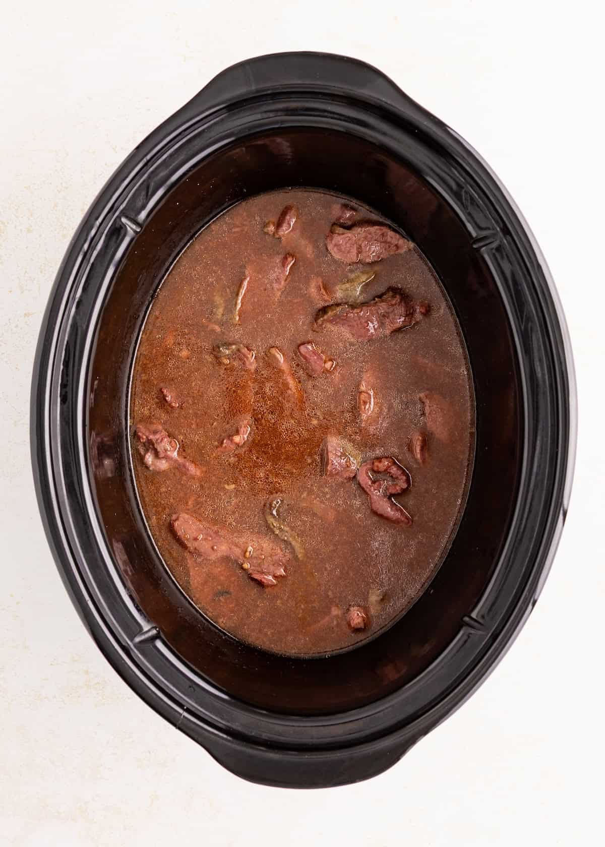 Sliced beef in brown sauce cooking inside a black slow cooker, viewed from above.
