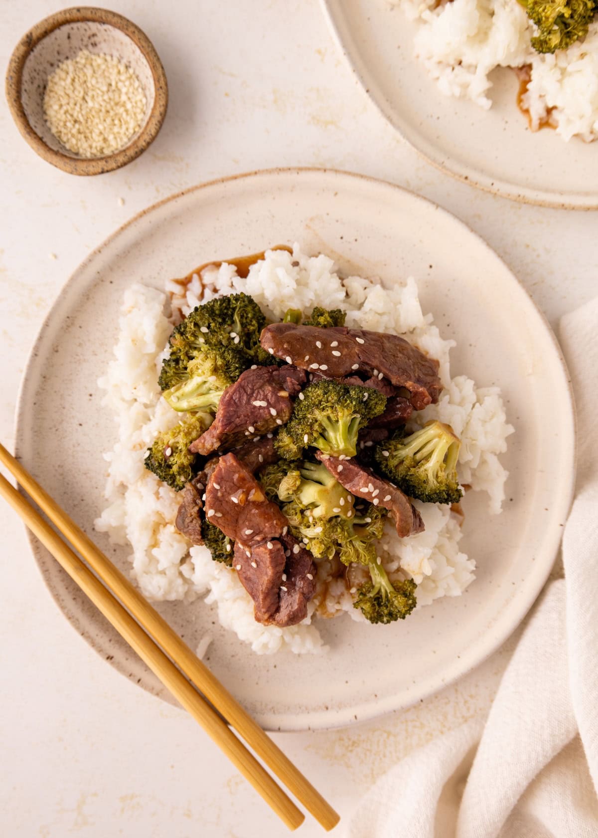 Plate of beef and broccoli stir-fry over white rice, with chopsticks and sesame seeds nearby.