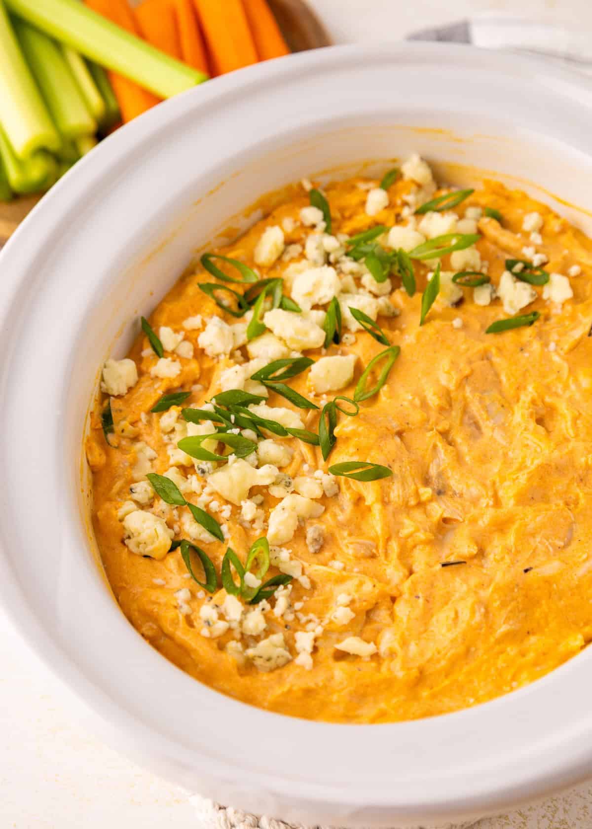 Buffalo chicken dip topped with green onions and cheese in a white dish, veggies in background.