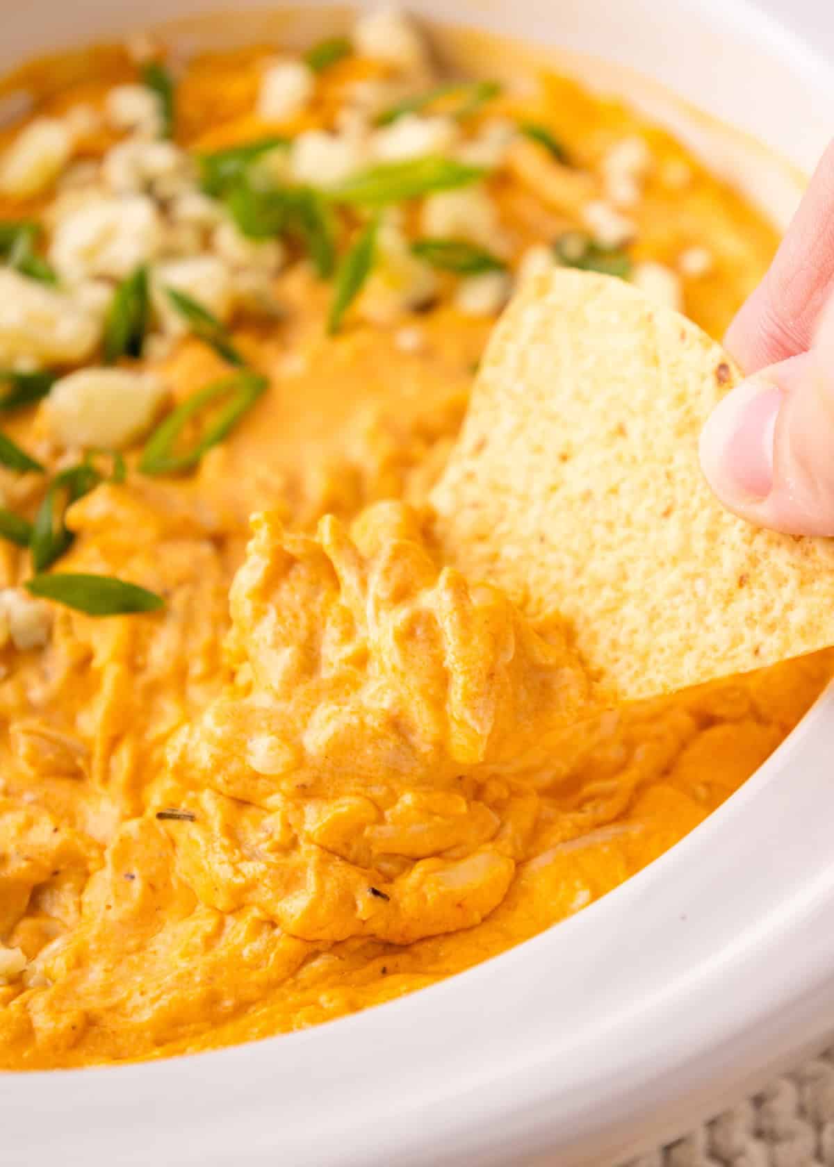 A hand dips a tortilla chip into a creamy, orange buffalo chicken dip in a white dish.