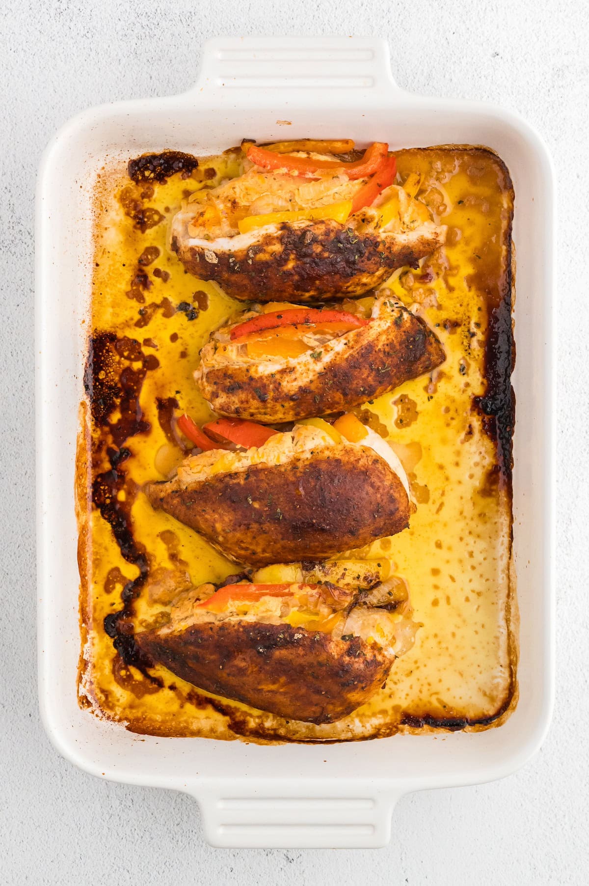 Four seasoned stuffed chicken breasts baked in a white rectangular dish with melted cheese and peppers.
