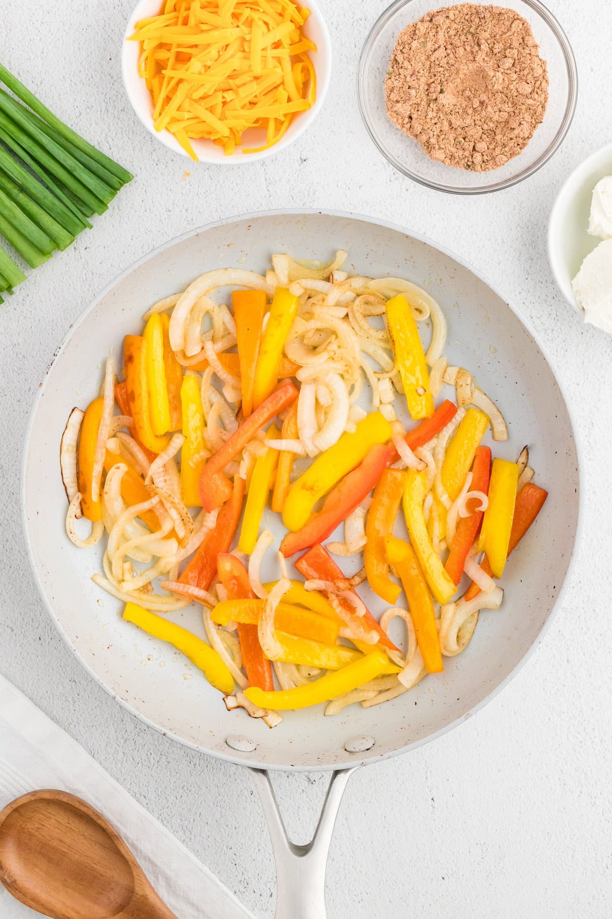 Sliced onions and bell peppers cooking in a pan, with cheese, spices, and scallions nearby.