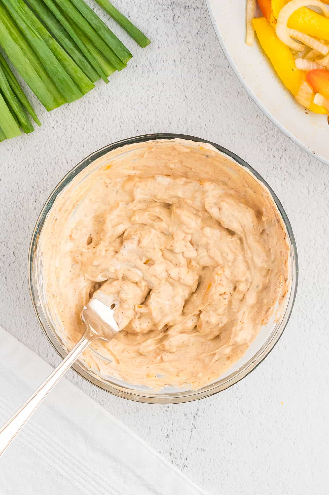 Bowl of creamy dip with a fork, next to green onions and sliced vegetables on a plate.