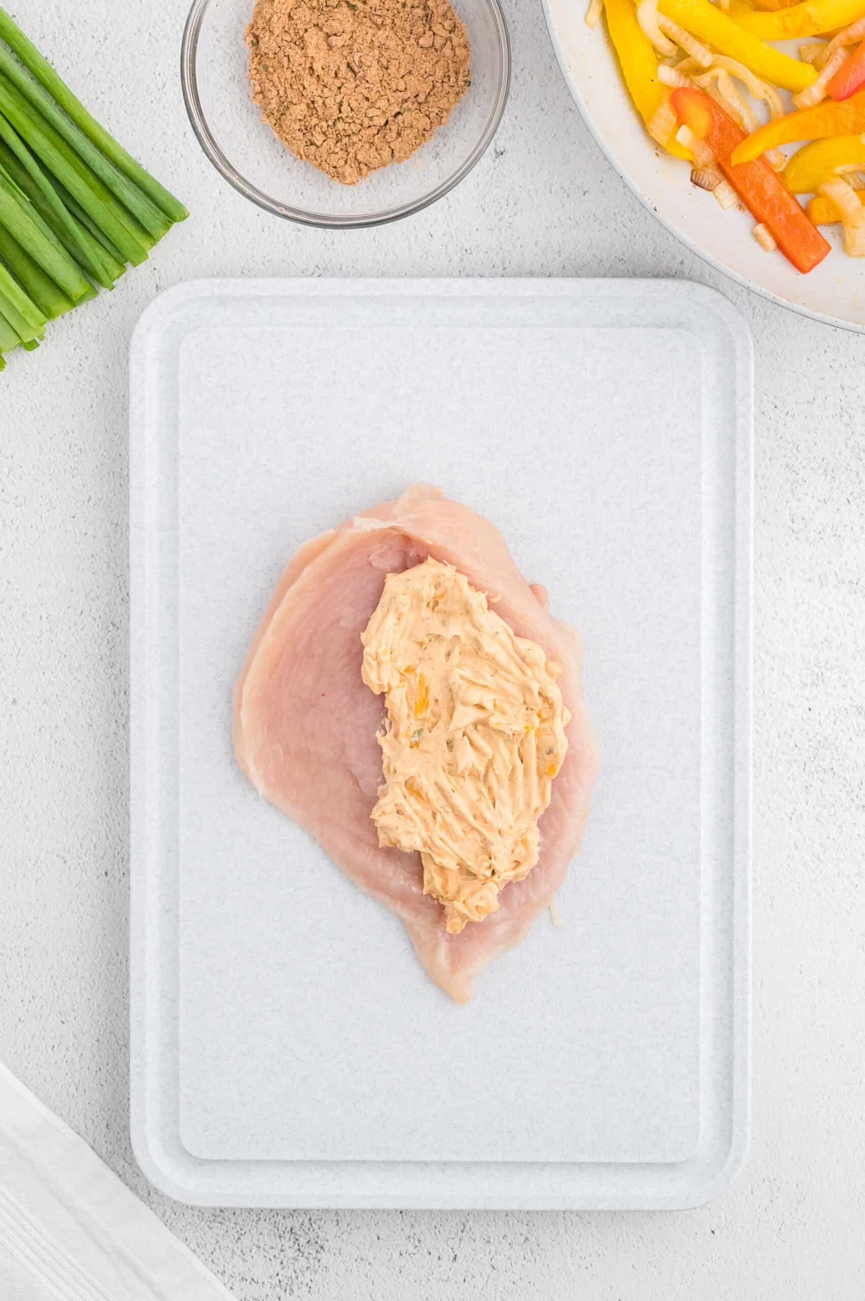 Raw chicken breast with cheese filling on a cutting board, veggies and seasonings nearby.
