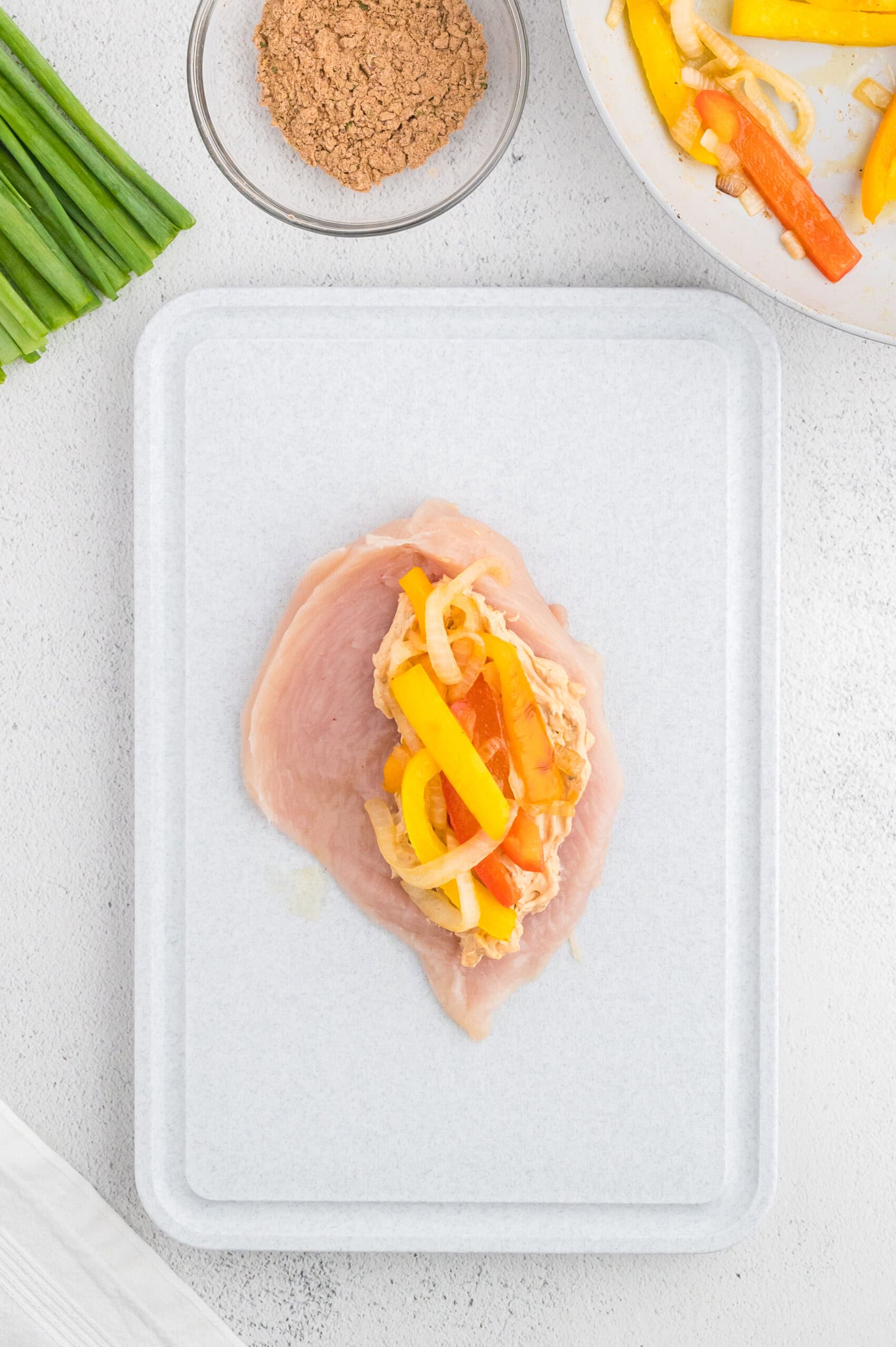 Raw chicken breast topped with sliced yellow and red bell peppers and onions on a cutting board.