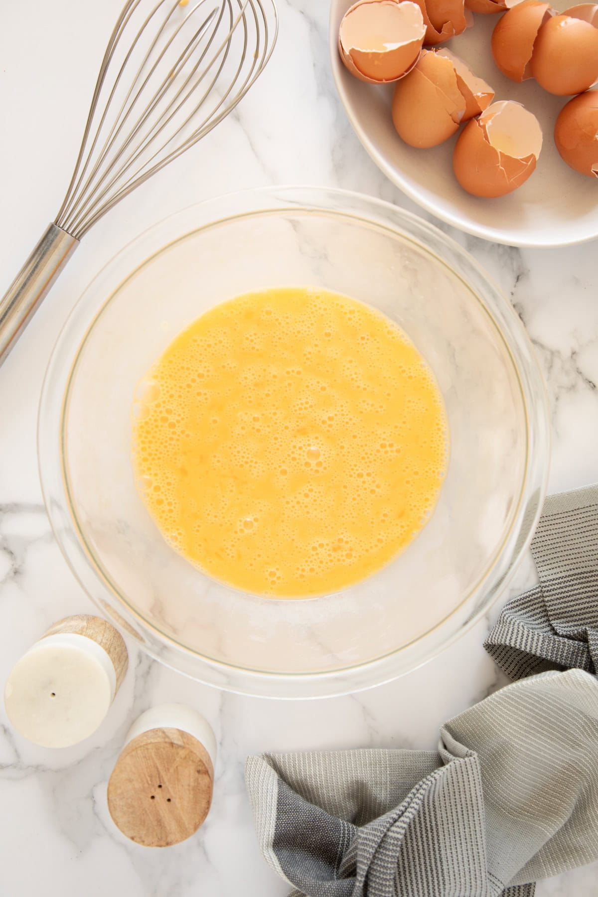 Beaten eggs in a glass bowl with egg shells, whisk, salt, pepper, and towel nearby.