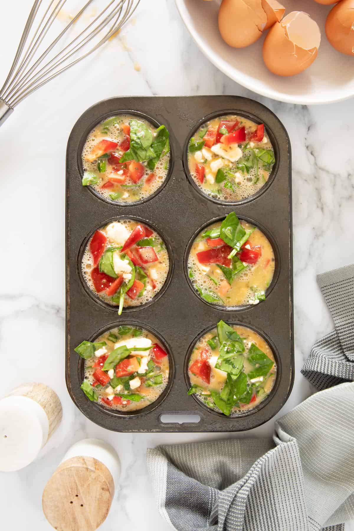 Muffin tin with six unbaked Feta Spinach Egg Muffins, surrounded by eggs and baking tools.