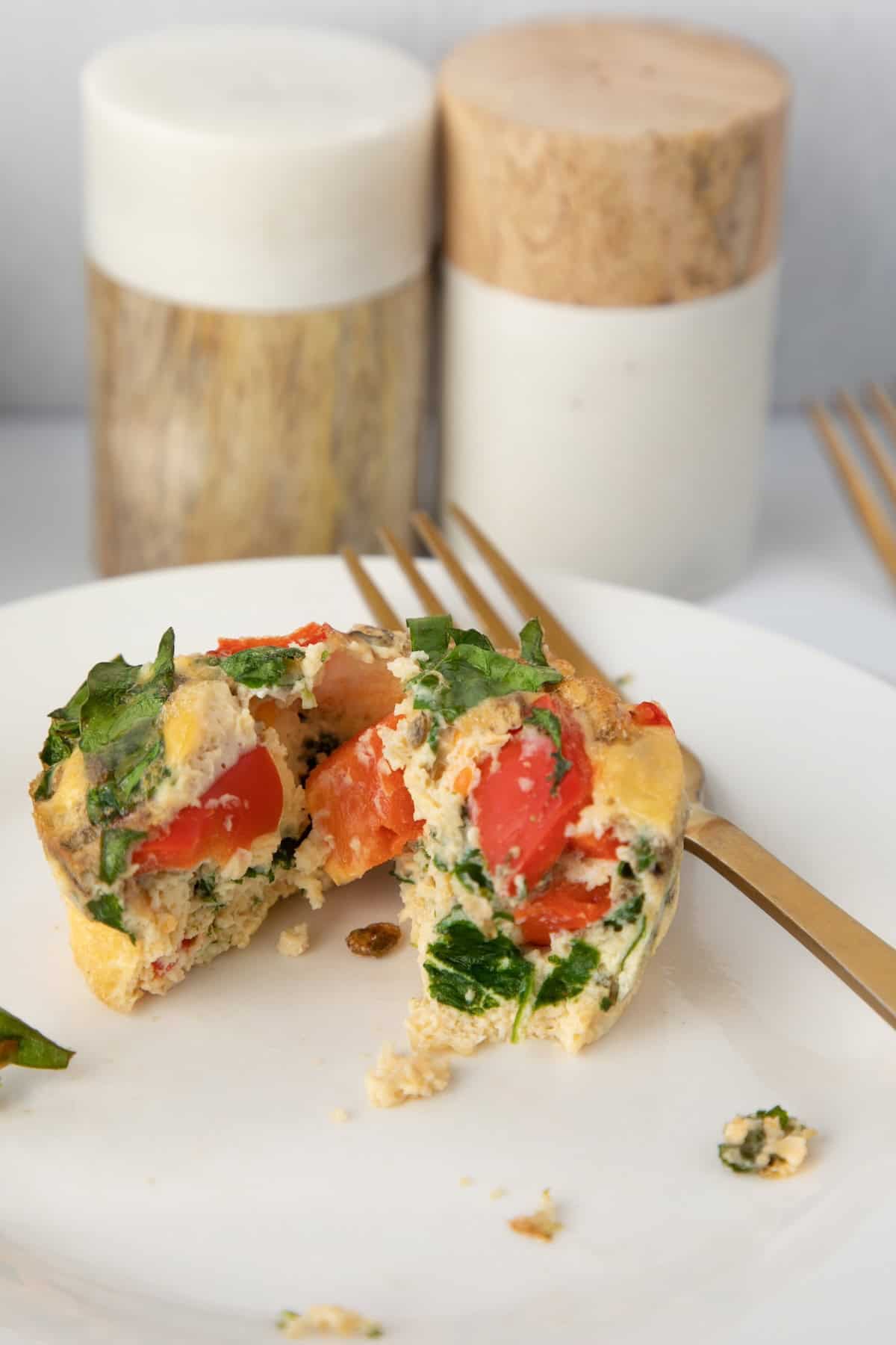 Egg muffin with spinach and tomatoes, cut open on a white plate with a gold fork.