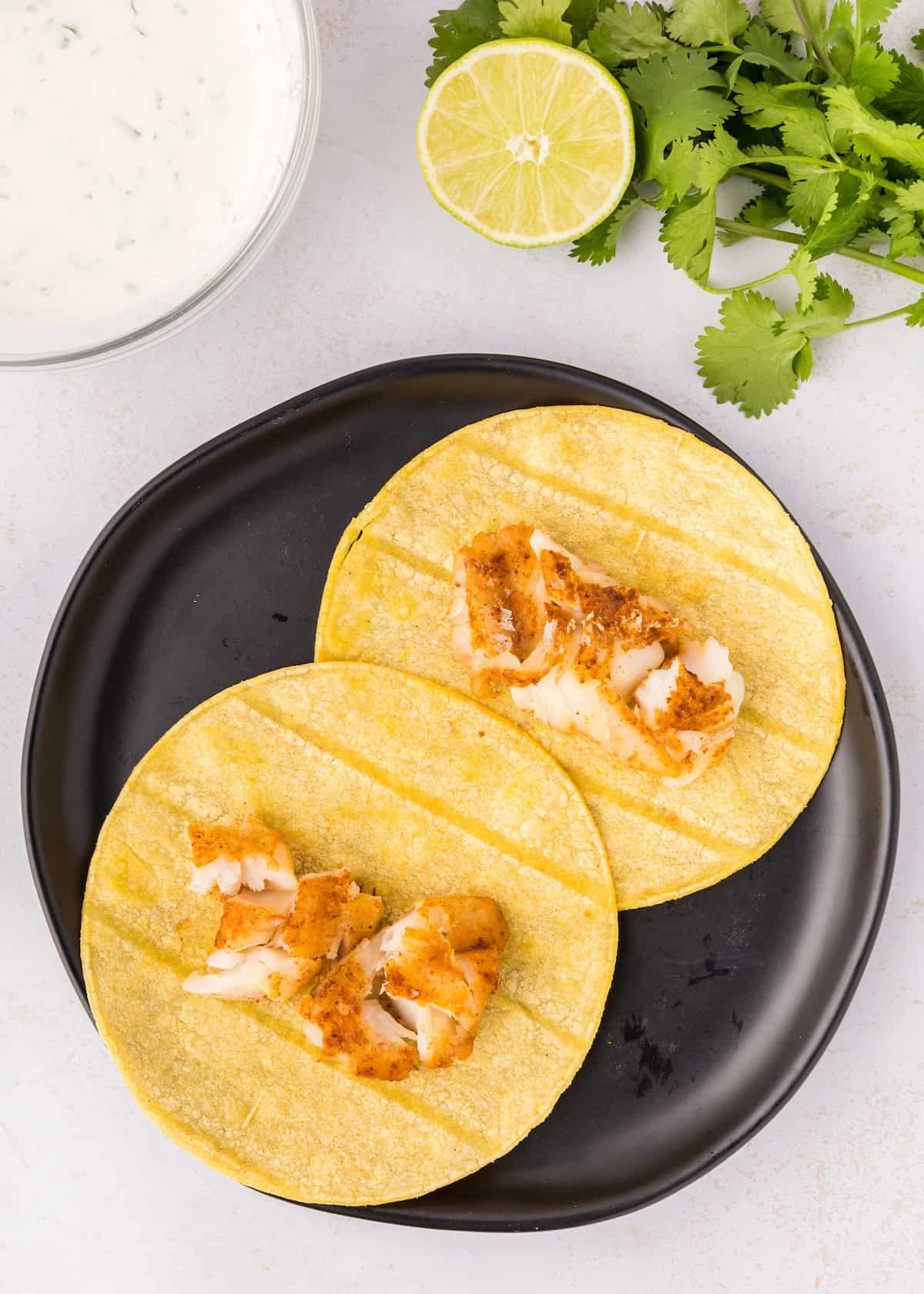 Two corn tortillas with grilled fish on a black plate, next to cilantro, lime, and sauce.