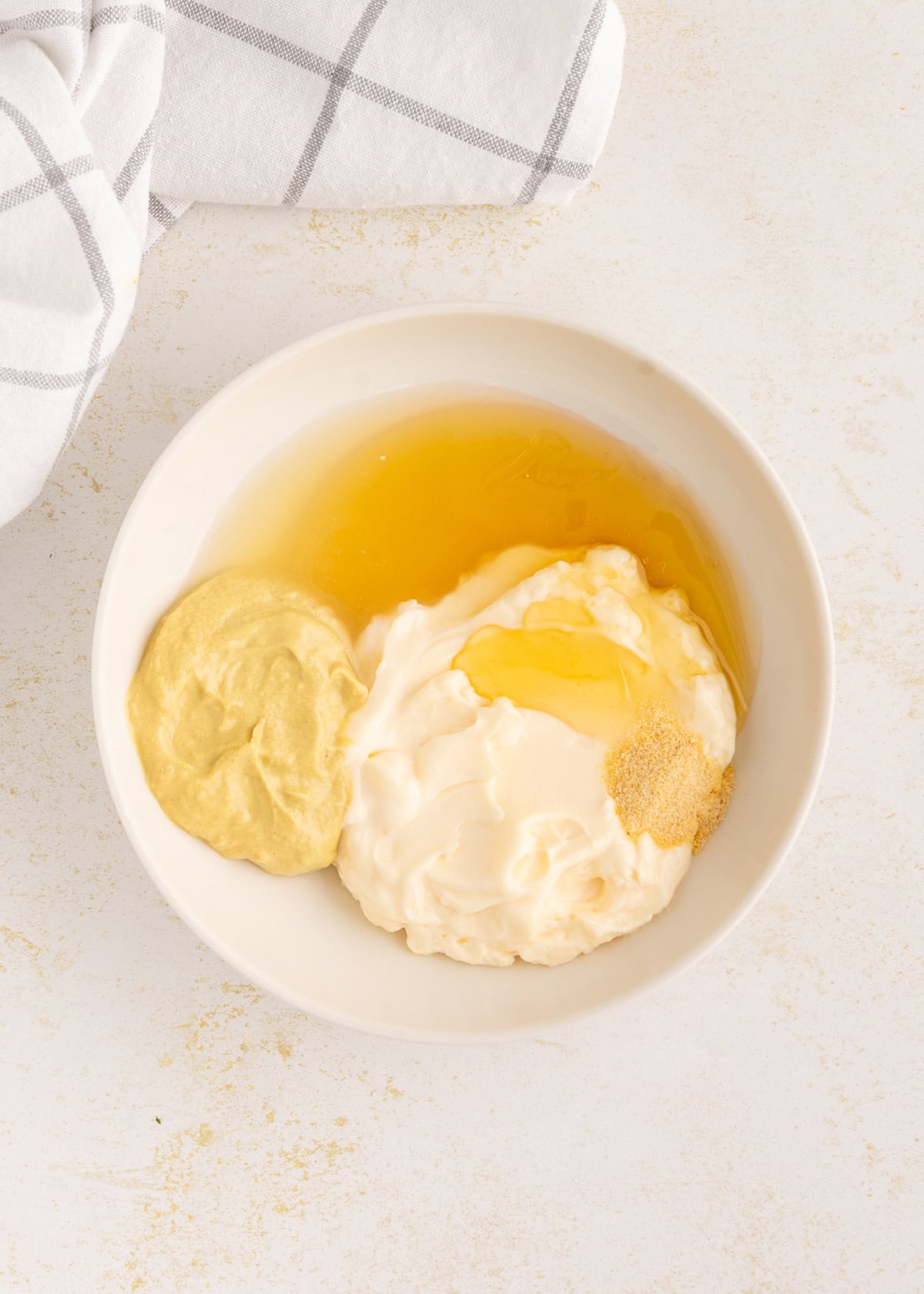 White bowl with Honey Mustard Dressing and seasoning on a white surface with a towel nearby.