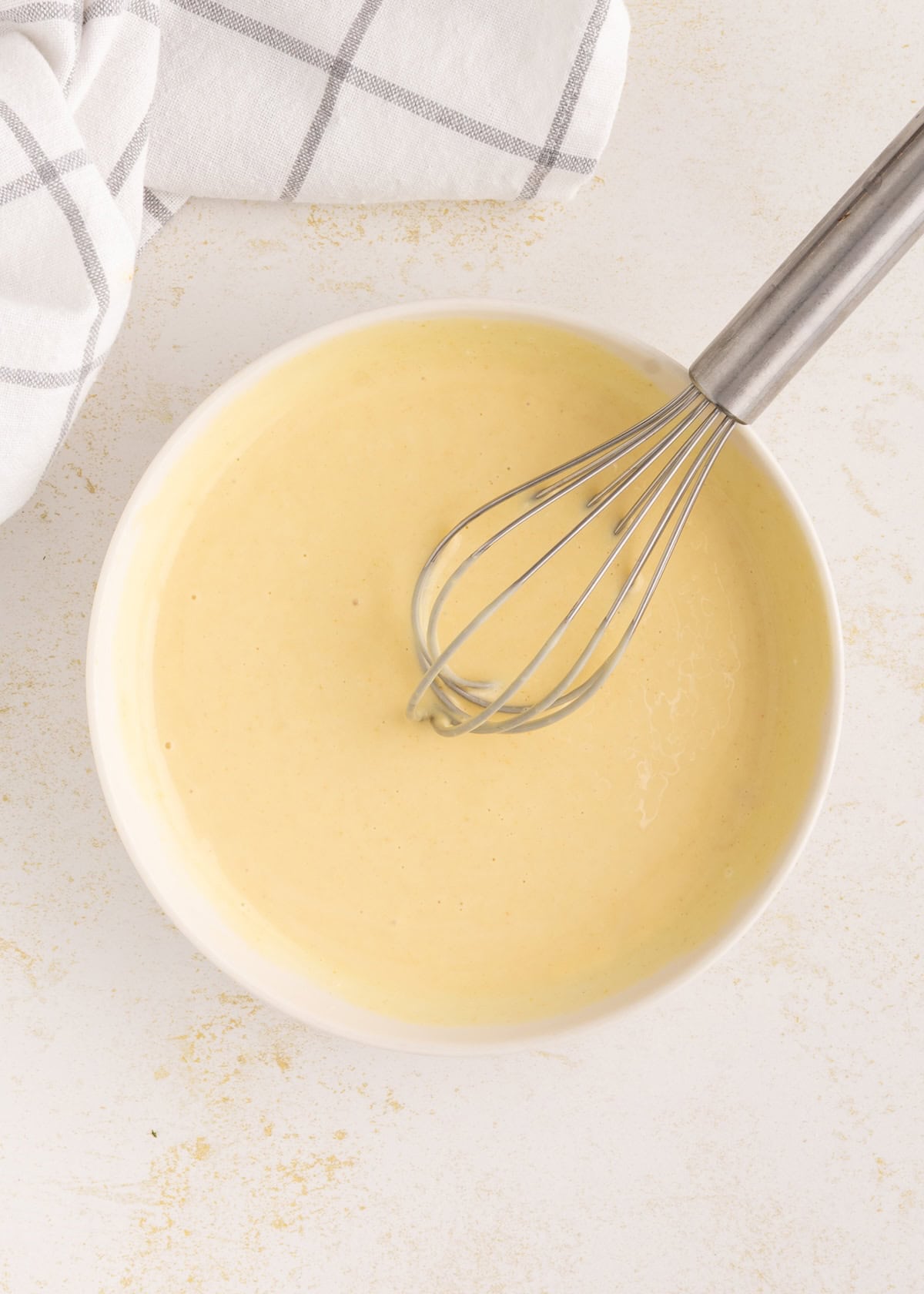 A metal whisk in a bowl of smooth yellow Honey Mustard Dressing beside a white checkered towel.