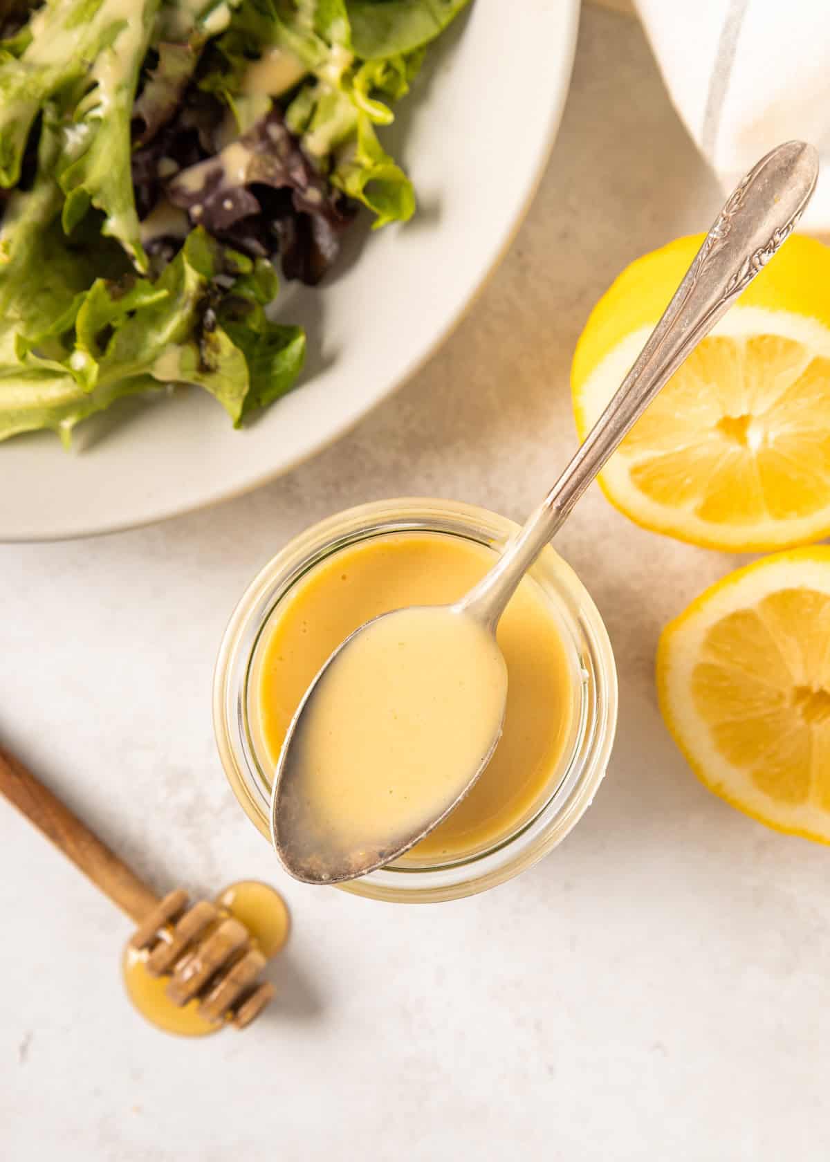 A spoon rests in a jar of Honey Mustard Dressing, with salad and lemon halves nearby.
