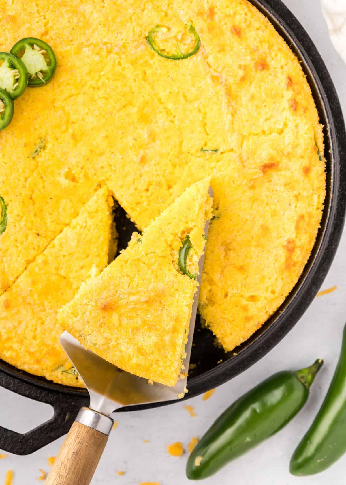 A slice of Jalapeño Cheddar Cornbread is lifted from a skillet, topped with fresh peppers.