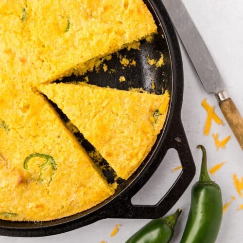 Jalapeño Cheddar Cornbread in a skillet, one slice cut, beside two jalapeños and a knife.