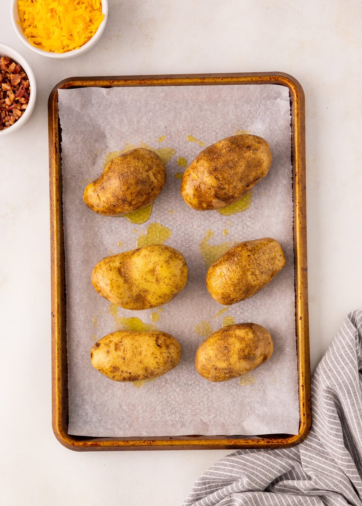 Six whole potatoes on a parchment-lined baking sheet, drizzled with oil, ready to bake.