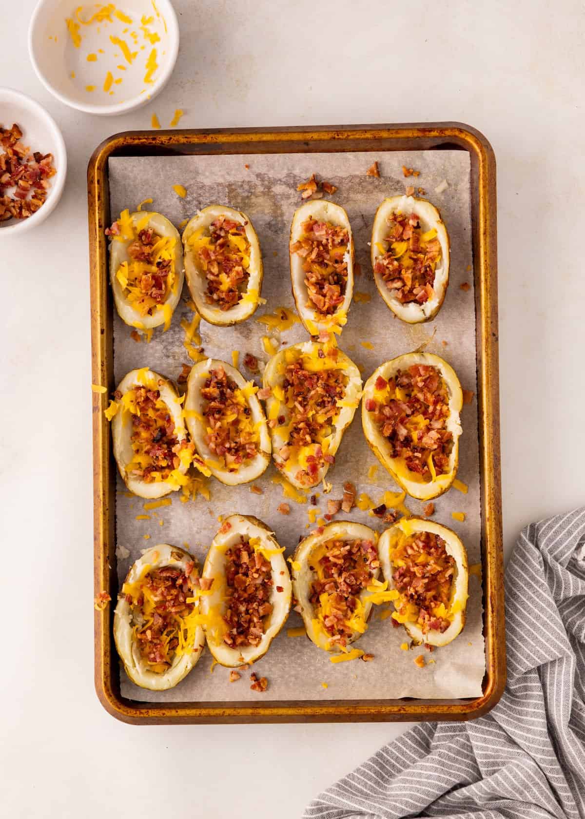 Potato skins on a baking sheet topped with cheese and bacon, with bowls of toppings nearby.