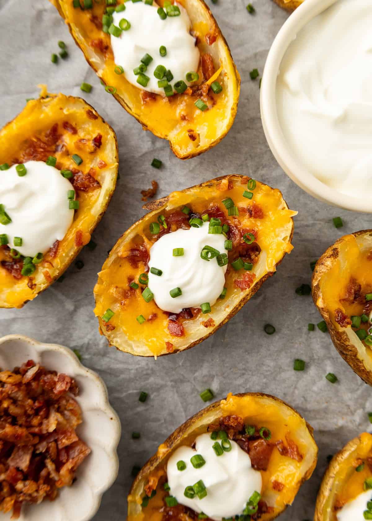 Baked potato skins with cheese, bacon, sour cream, and chives on parchment paper.