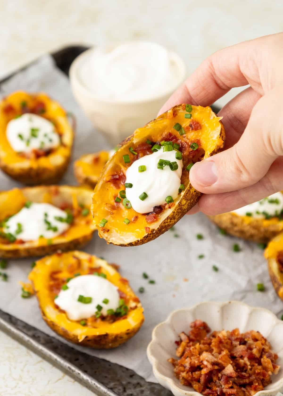 A hand holds a loaded potato skin topped with cheese, sour cream, bacon, and chives.