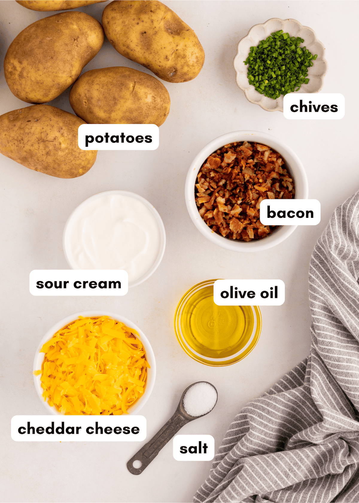 Top view of potatoes, chives, bacon, sour cream, olive oil, cheddar cheese, and salt on a table.