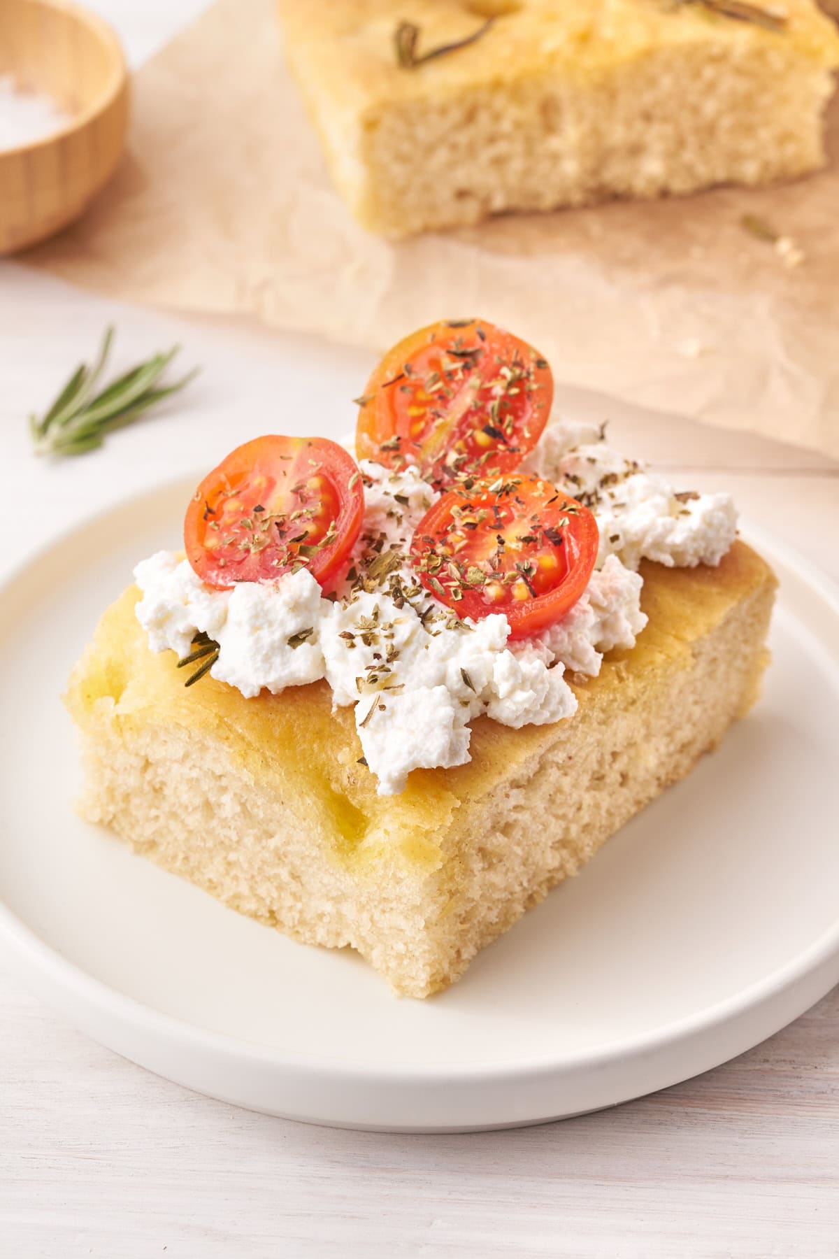 A slice of focaccia topped with ricotta, cherry tomatoes, and herbs on a white plate.