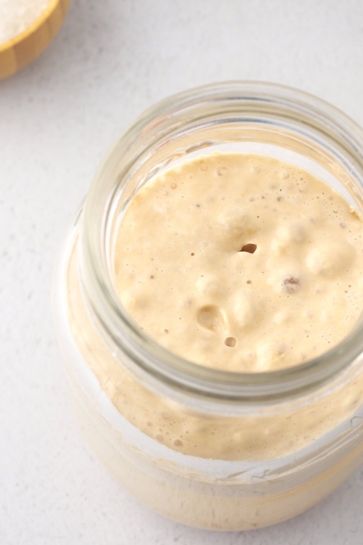 A glass jar filled with bubbly, active sourdough starter on a light surface.