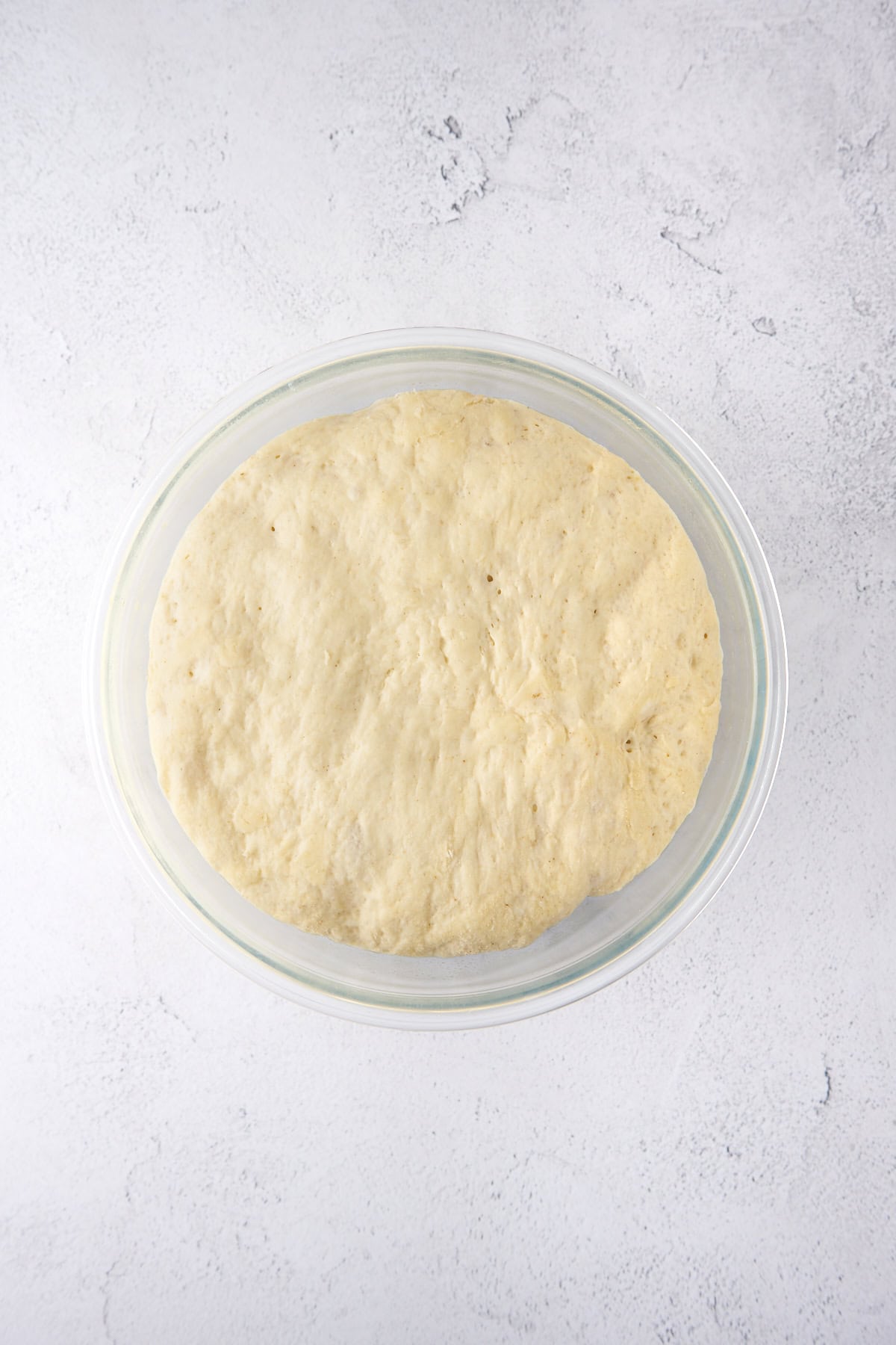 A glass bowl filled with risen dough on a light, textured surface.
