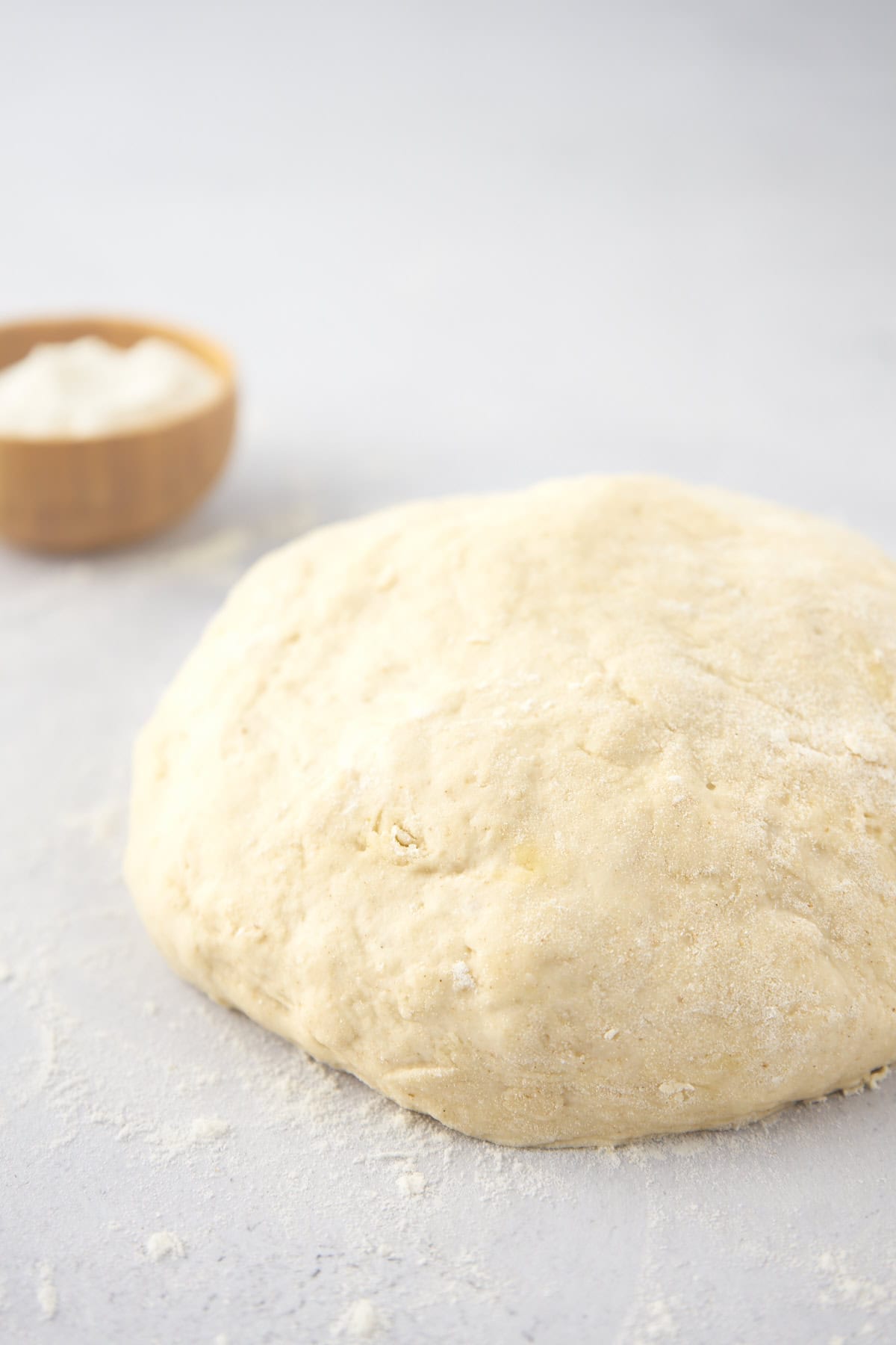 A ball of dough on a floured surface with a small bowl of flour in the background.