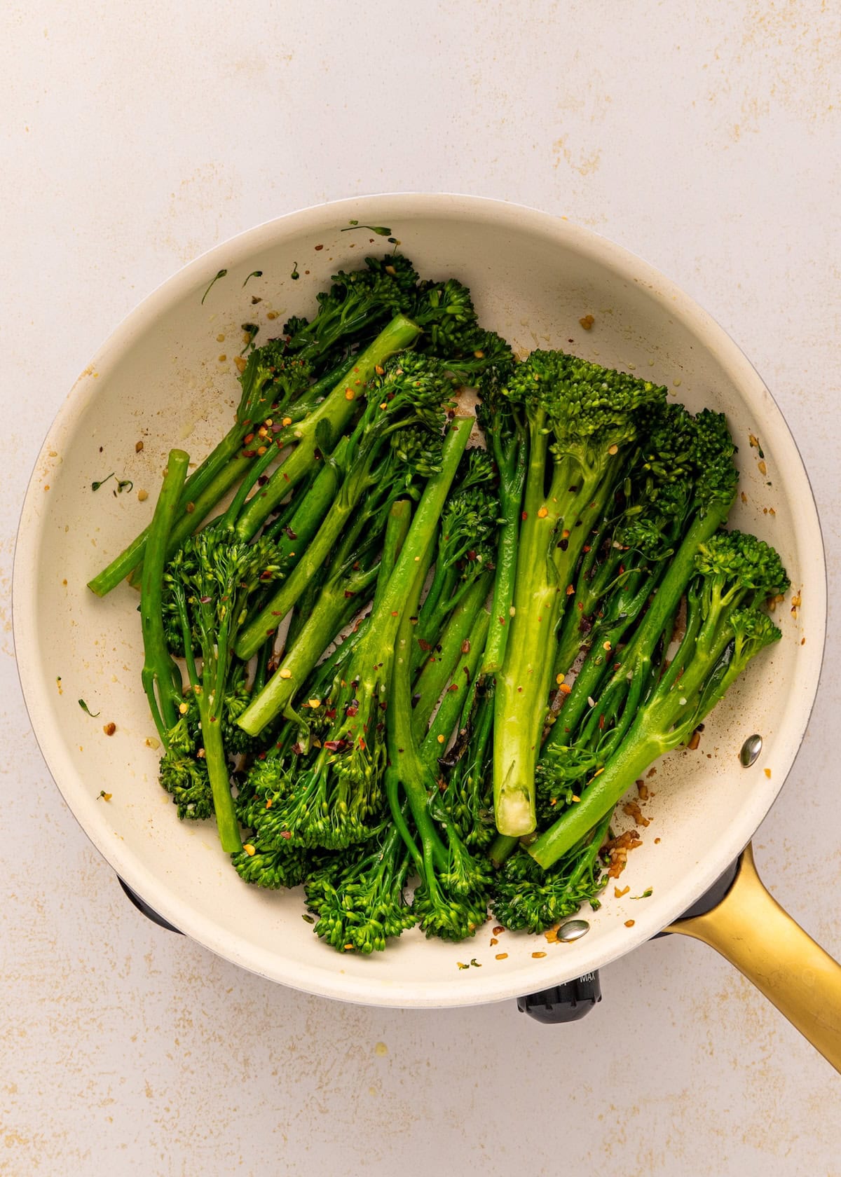Broccolini sautéing in a light-colored pan with seasonings on a light background.