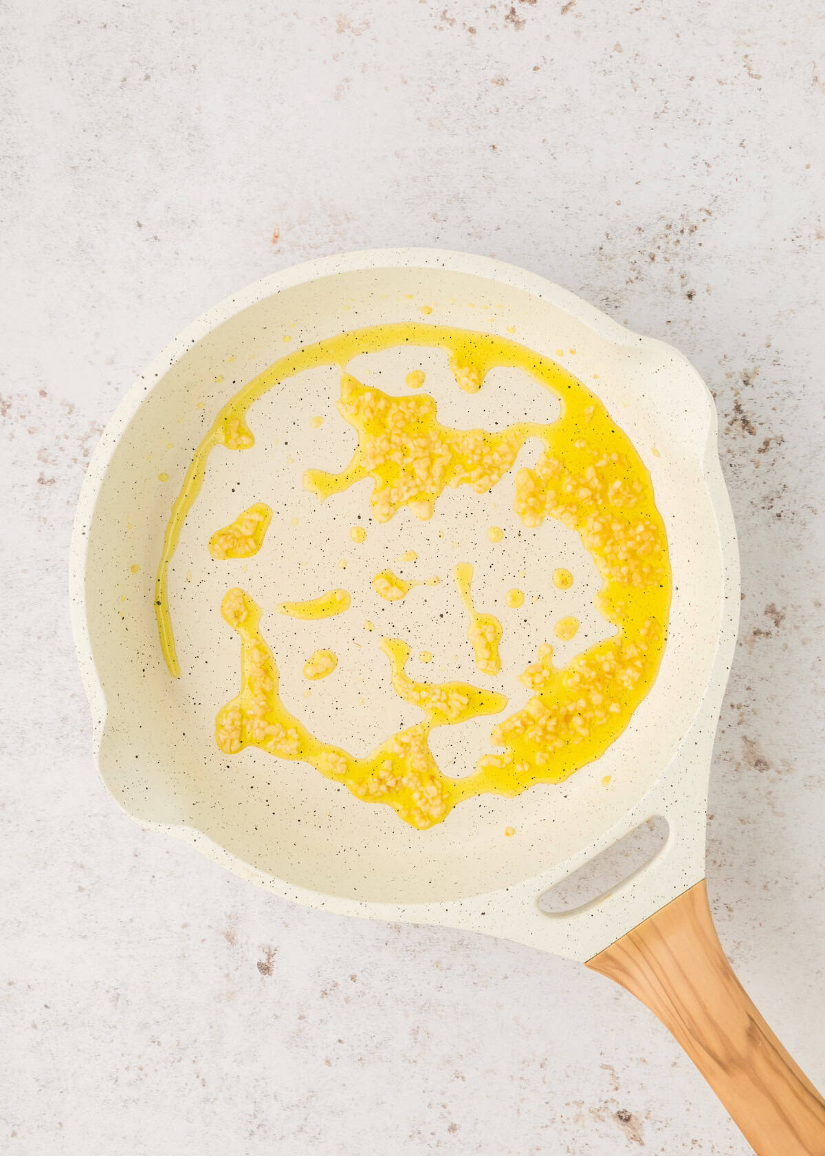 White skillet with melted butter and minced garlic on a light countertop, seen from above.